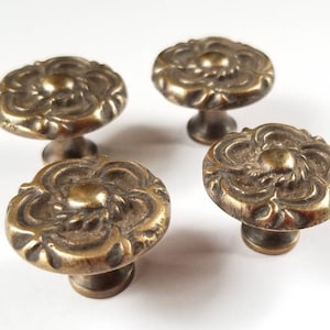 Peut inclure: Quatre boutons de meuble en laiton antique avec un motif floral. Chaque bouton présente un motif floral en relief détaillé sur une face ronde, monté sur une base cylindrique courte. Les boutons ont une finition en laiton vieilli et chaleureux.