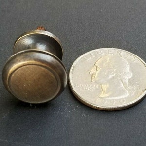 May include: A small, round, brass cabinet knob with a raised edge and a screw on the back. The knob is about the size of a US quarter.