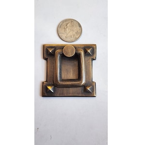 Pode incluir: Uma puxador de armário quadrado de latão antigo com uma alça de anel e detalhes decorativos. O puxador tem um acabamento desgastado e um design ligeiramente elevado.