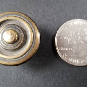 May include: A brass cabinet knob with a concentric circle design. The knob has a gold-colored center and is about the size of a US quarter.