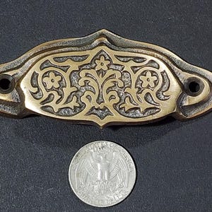 Puede incluir: Un tirador de cajón de latón con un diseño floral ornamentado. El tirador es de color dorado claro y tiene un acabado ligeramente envejecido.