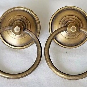 Op de afbeelding: Twee antieke messing kastdeurgrepen met een ringvormige handgreep. De grepen hebben een ronde, vlakke basis met concentrische ringen.