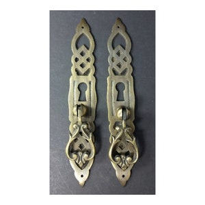 May include: Two antique brass cabinet door pulls with ornate designs and keyhole cutouts. The pulls have a decorative loop handle.
