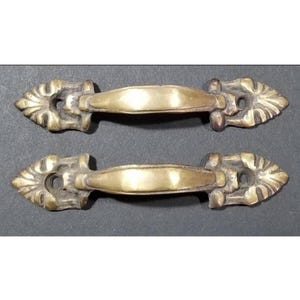 May include: Two antique brass cabinet door pulls with ornate designs. The pulls have a simple, rectangular shape with a slight curve in the middle.