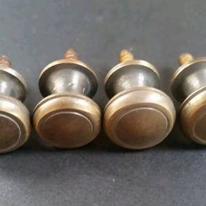 May include: Four brass cabinet knobs with a round, slightly raised top and a smaller, round base. The knobs have a screw on the bottom for attaching to a surface.