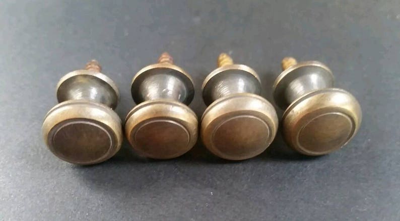 May include: Four brass cabinet knobs with a round, slightly raised top and a smaller, round base. The knobs have a screw on the bottom for attaching to a surface.