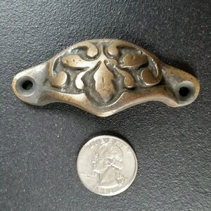 Peut inclure: Une poignée de tiroir en laiton avec un motif floral décoratif. La poignée est à peu près de la taille d'une pièce de 20 centimes.