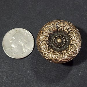 Op de afbeelding: Een gouden metalen knop met een bloemmotief. De knop heeft een verhoogd midden met een kleinere cirkel in het midden.