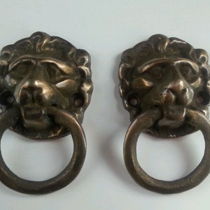 Könnte beinhalten: Zwei antike Löwenkopf-Schubladengriffe mit einem Ringgriff. Die Griffe sind aus Metall und haben eine Bronze-Oberfläche.