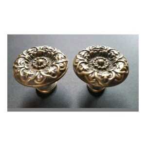 May include: A pair of antique bronze cabinet knobs featuring a floral design. The knobs have a round, ornate face with detailed patterns and a short, cylindrical base. The finish has a warm, aged appearance.