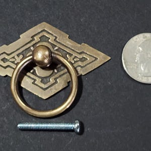 May include: A brass cabinet pull with a diamond-shaped plate and a ring handle. The plate has an intricate design and the handle is smooth. The pull comes with a screw for installation.