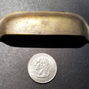 May include: A brass cabinet pull with a curved top and rounded edges. The pull is about the size of a US quarter.