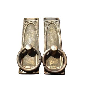 May include: Two antique brass door pulls with a ring handle. The pulls are rectangular with an arched top and a decorative design.