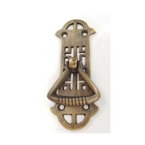 May include: A brass cabinet pull with a decorative, geometric design. The pull has a loop handle and is attached to a rectangular plate.