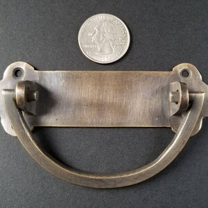 Op de afbeelding: Antiek messing deurbeslag met een rechthoekige plaat en een gebogen handvat. Het beslag is ongeveer 10 cm lang en 5 cm breed.