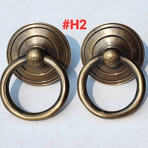 Może przedstawiać: Dwa antyczne mosiężne uchwyty do szafek z uchwytem w kształcie pierścienia. Uchwyty mają okrągłą podstawę z koncentrycznymi pierścieniami.