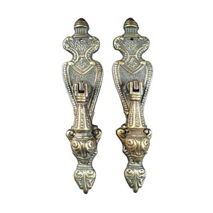 May include: A pair of ornate brass door pulls with intricate designs. The pulls have a  distinctive, almost animal-like, shape at the bottom.