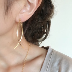 May include: Gold-colored, long, curved earrings with a delicate chain necklace. The earrings have a slender, elongated design, creating a graceful, flowing appearance. The jewelry is set against a neutral background, highlighting the warm tones of the metal.