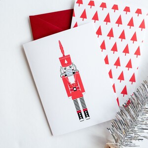 May include: A white card with a red and black illustration of a nutcracker wearing a face mask. The card is surrounded by a red envelope and a silver tinsel garland.