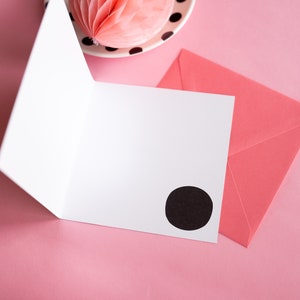 Op de afbeelding: Een open witte wenskaart met een zwarte cirkel op de voorkant, naast een roze envelop. Een roze honingraatdecoratie en een bord met zwarte stippen zijn op de achtergrond, alles op een roze oppervlak.