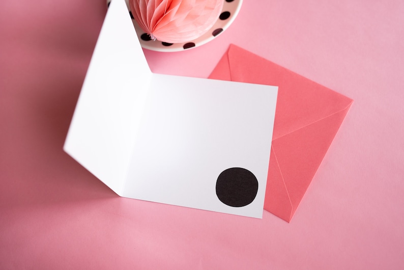 Op de afbeelding: Een open witte wenskaart met een zwarte cirkel op de voorkant, naast een roze envelop. Een roze honingraatdecoratie en een bord met zwarte stippen zijn op de achtergrond, alles op een roze oppervlak.