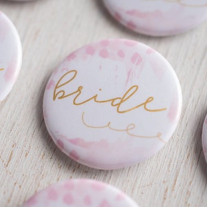 May include: A set of round, white button pins with pink watercolor designs. The pins say "bride" in gold lettering.
