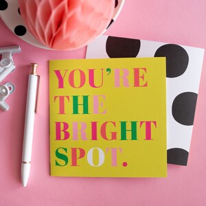 Op de afbeelding: Een vrolijke compositie met een gele kaart met de tekst "YOU'RE THE BRIGHT SPOT" in roze en groen. Een witte pen, paperclips en een kaart met stippen zijn gerangschikt op een roze achtergrond, samen met een decoratieve honingraatbal.