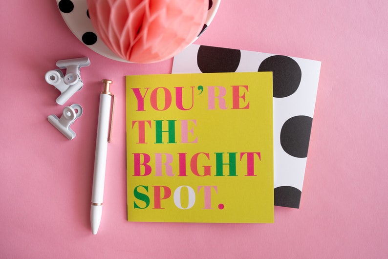 Op de afbeelding: Een vrolijke compositie met een gele kaart met de tekst "YOU'RE THE BRIGHT SPOT" in roze en groen. Een witte pen, paperclips en een kaart met stippen zijn gerangschikt op een roze achtergrond, samen met een decoratieve honingraatbal.