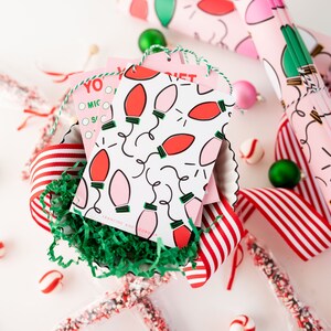 May include: Pink and red gift wrap with a pattern of light bulbs and a white background. The gift wrap is rolled up and tied with a green ribbon. There are also some red and white striped ribbon, candy canes, and ornaments in the image.