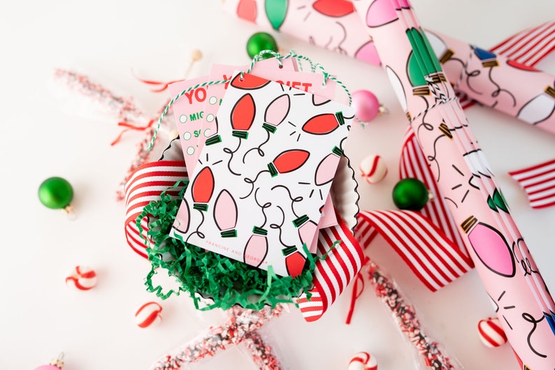 May include: Pink and red gift wrap with a pattern of light bulbs and a white background. The gift wrap is rolled up and tied with a green ribbon. There are also some red and white striped ribbon, candy canes, and ornaments in the image.