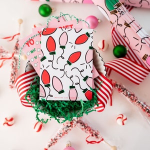 May include: A close-up of a pink and white gift tag with a red and pink lightbulb pattern. The tag is in a bowl with green shredded paper and red and white striped ribbon. The text on the tag reads "Francine and Robert".
