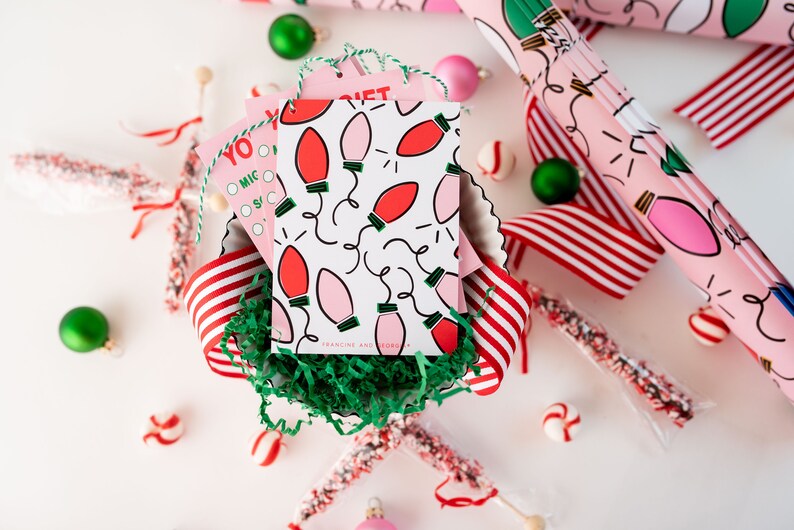 May include: A close-up of a pink and white gift tag with a red and pink lightbulb pattern. The tag is in a bowl with green shredded paper and red and white striped ribbon. The text on the tag reads "Francine and Robert".