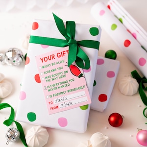 May include: A white gift box with pink and green polka dots wrapped with a green ribbon. A pink gift tag with the text "YOUR GIFT" and a checklist of options is attached to the box. The text on the tag reads: "MIGHT BE ALIVE, SCREAMS YOU, WAS BOUGHT ON THE WAY HERE, IS EVERYTHING YOU NEVER WANTED, IS POSSIBLY PERISHABLE". The tag also has a space to write "TO" and "FROM". The text on the tag reads: "To Hallis, FROM V Santa."