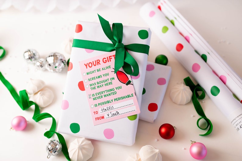 May include: A white gift box with pink and green polka dots wrapped with a green ribbon. A pink gift tag with the text "YOUR GIFT" and a checklist of options is attached to the box. The text on the tag reads: "MIGHT BE ALIVE, SCREAMS YOU, WAS BOUGHT ON THE WAY HERE, IS EVERYTHING YOU NEVER WANTED, IS POSSIBLY PERISHABLE". The tag also has a space to write "TO" and "FROM". The text on the tag reads: "To Hallis, FROM V Santa."