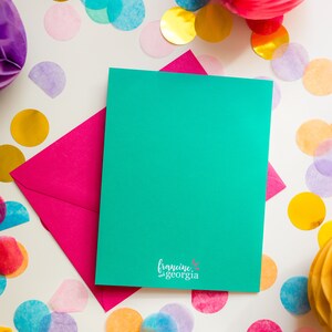 May include: A teal greeting card and a pink envelope are displayed on a white surface, surrounded by colorful confetti and paper decorations. The card has the text "francine and georgia" at the bottom.