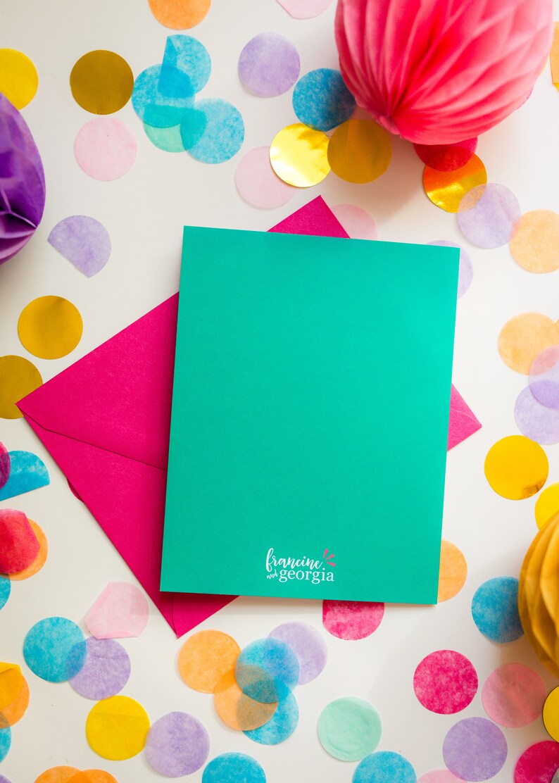 May include: A teal greeting card and a pink envelope are displayed on a white surface, surrounded by colorful confetti and paper decorations. The card has the text "francine and georgia" at the bottom.