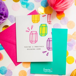 May include: Greeting card with the text "YOU'RE A PRECIOUS FUCKING GEM" and colorful gem illustrations. The card is on a white surface with a pink envelope, teal card, and confetti.