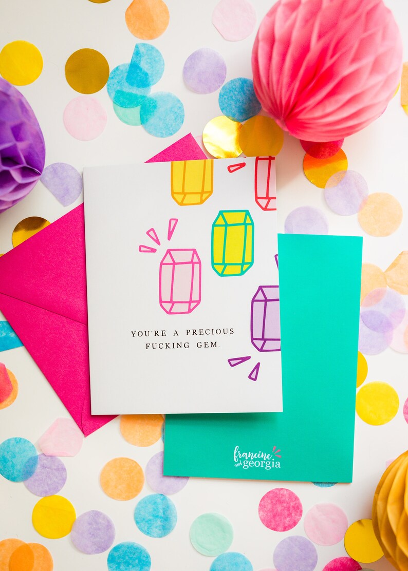 May include: Greeting card with the text "YOU'RE A PRECIOUS FUCKING GEM" and colorful gem illustrations. The card is on a white surface with a pink envelope, teal card, and confetti.