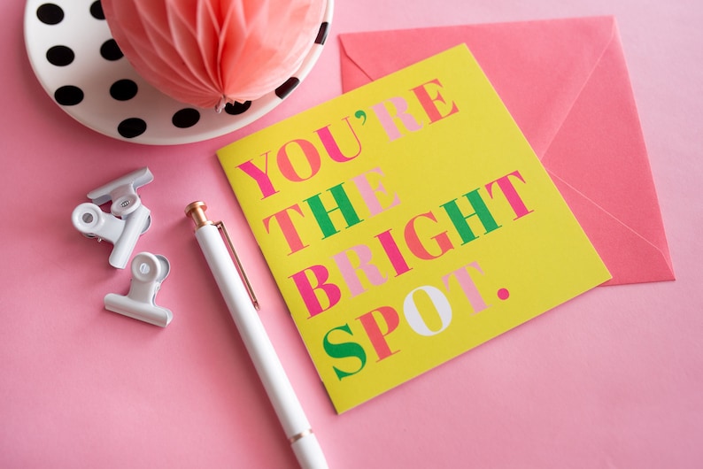 Op de afbeelding: Een gele wenskaart met de tekst "YOU'RE THE BRIGHT SPOT" in roze, groen en wit. Een roze envelop, witte pen en zilveren paperclips liggen op een roze ondergrond. Een roze honingraatdecoratie en een bord met zwarte en witte stippen staan op de achtergrond.