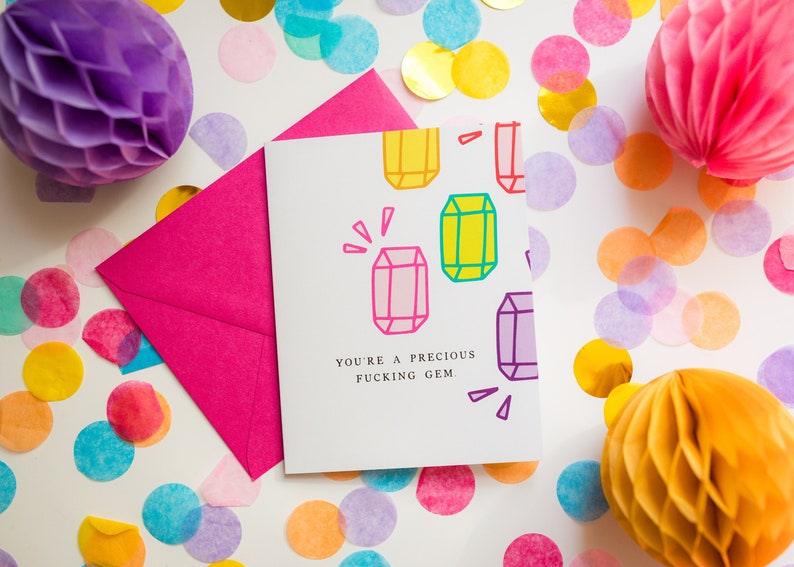 May include: A greeting card with the text "YOU'RE A PRECIOUS FUCKING GEM" and colorful gem illustrations. A bright pink envelope and confetti surround the card, with honeycomb paper decorations in purple, pink, and orange.