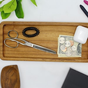 May include: A wooden tray with a pair of scissors, a hair tie, a small stack of US coins, a dollar bill, and a white case. The tray is on a white surface.