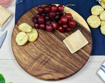 May include: A round wooden serving board with a dark brown wood grain. The board is topped with a cluster of red grapes, a small piece of cheese, and several crackers.