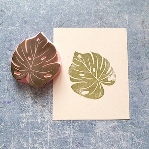 May include: A brown rubber stamp with a detailed monstera leaf design. The stamp is on a white card with a green monstera leaf print.