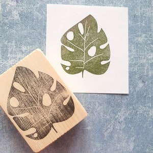 May include: A wooden stamp with a black monstera leaf design next to a print of the same leaf in green ink on white paper. The stamp is rectangular and the leaf has several holes.