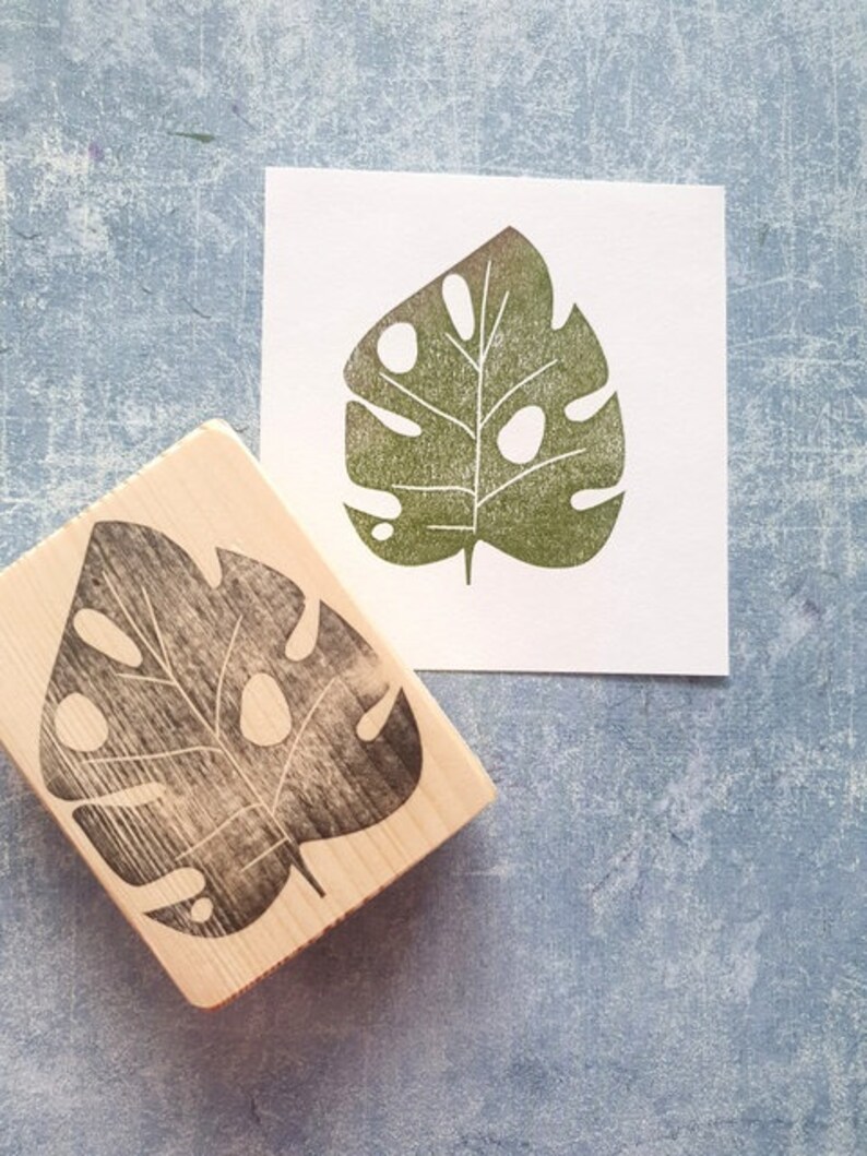 May include: A wooden stamp with a black monstera leaf design next to a print of the same leaf in green ink on white paper. The stamp is rectangular and the leaf has several holes.