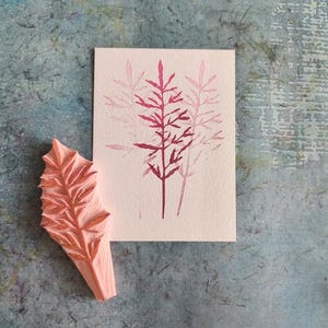 Könnte beinhalten: Ein rosa Gummistempel mit einem Blattdesign neben einer Karte mit einem passenden Blattdruck in Rosatönen. Der Stempel und die Karte befinden sich auf einer strukturierten blau-grauen Oberfläche.