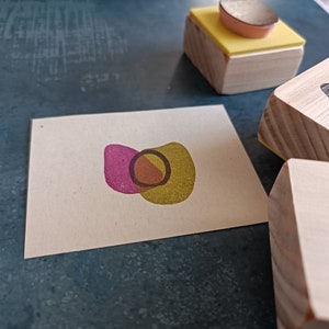 May include: A set of three wooden stamps with different abstract designs. The stamps are made of natural wood and have a light brown color. The designs are a pink and yellow overlapping shape, a black leaf shape, and a black circle.