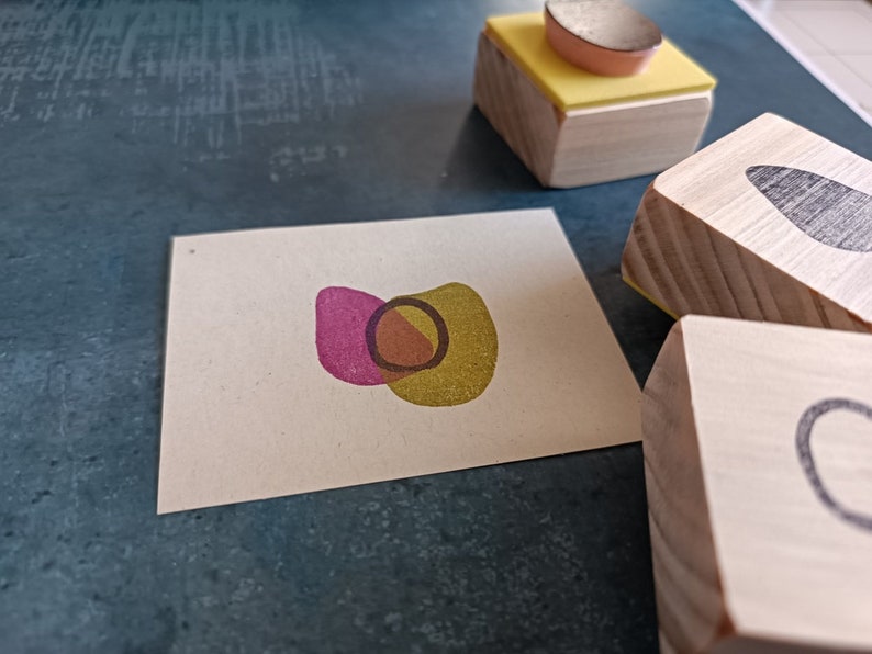 May include: A set of three wooden stamps with different abstract designs. The stamps are made of natural wood and have a light brown color. The designs are a pink and yellow overlapping shape, a black leaf shape, and a black circle.