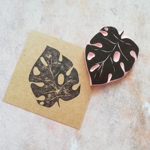 May include: A black and pink leaf-shaped stamp and a brown paper with a black leaf print. The stamp has a detailed design with cut-out sections. The print on the paper is a mirror image of the stamp, showcasing the intricate leaf pattern.
