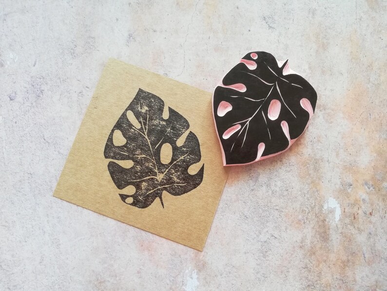 May include: A black and pink leaf-shaped stamp and a brown paper with a black leaf print. The stamp has a detailed design with cut-out sections. The print on the paper is a mirror image of the stamp, showcasing the intricate leaf pattern.